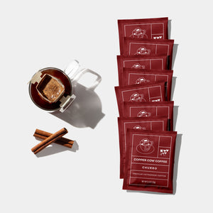 Multiple pre-filled single-serve pour over filters with a coffee blend, a cup of brewed coffee with cinnamon sticks, and the brand name 'Copper Cow Coffee' visible.