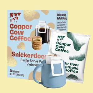 Copper Cow Coffee Snickerdoodle single-serve packet shown with its retail box and a blue ceramic mug holding one of the packets, on a pale yellow background.